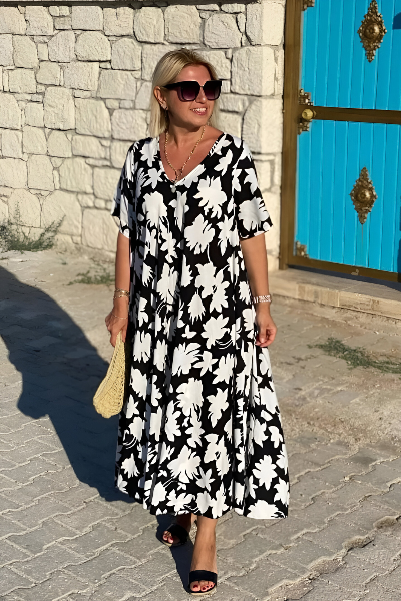 White - A woman wearing a black and white botanical leaf print maxi dress with a V-neckline and short sleeves, walking outdoors on a sunny day.