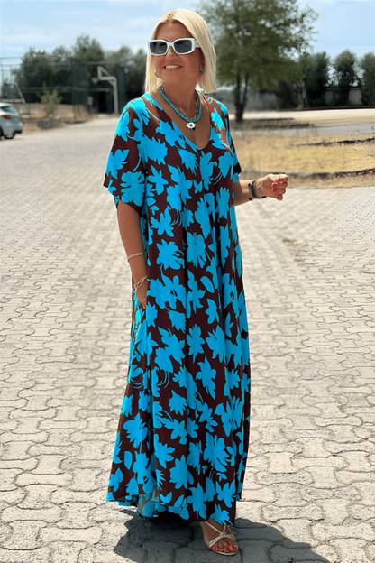Blue - A woman wearing a black and white botanical leaf print maxi dress with a V-neckline and short sleeves, walking outdoors on a sunny day.