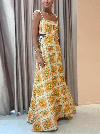 A woman wearing an orange and yellow lemon print maxi dress with side cutouts, tie-shoulder straps, and a sweeping skirt, standing against a textured plaster wall.