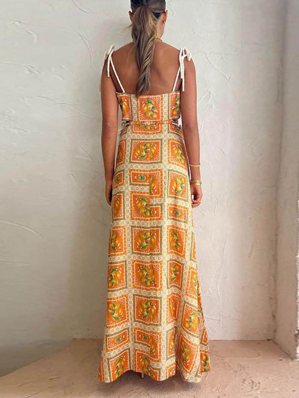 A woman wearing an orange and yellow lemon print maxi dress with side cutouts, tie-shoulder straps, and a sweeping skirt, standing against a textured plaster wall.