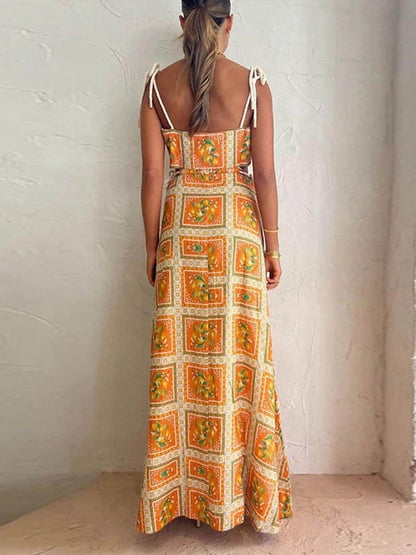 A woman wearing an orange and yellow lemon print maxi dress with side cutouts, tie-shoulder straps, and a sweeping skirt, standing against a textured plaster wall.
