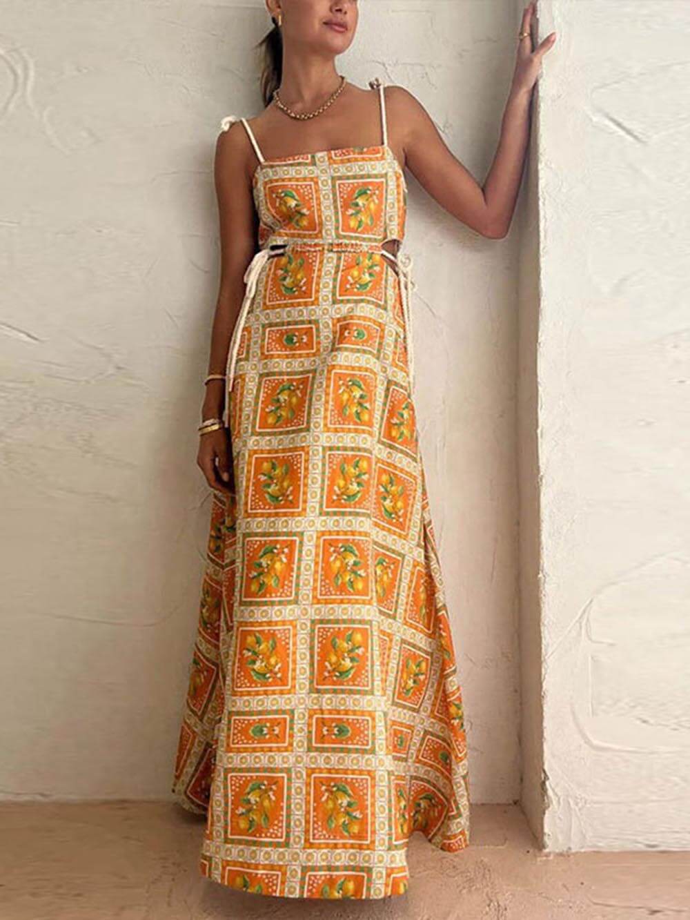 Yellow - A woman wearing an orange and yellow lemon print maxi dress with side cutouts, tie-shoulder straps, and a sweeping skirt, standing against a textured plaster wall.