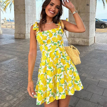 Yellow - Woman wearing a white mini dress with an all-over yellow lemon and green leaf print, featuring tie shoulder straps and a square neckline.