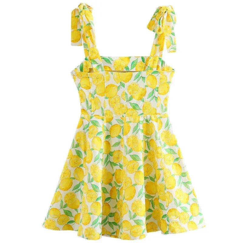 Yellow - Woman wearing a white mini dress with an all-over yellow lemon and green leaf print, featuring tie shoulder straps and a square neckline.