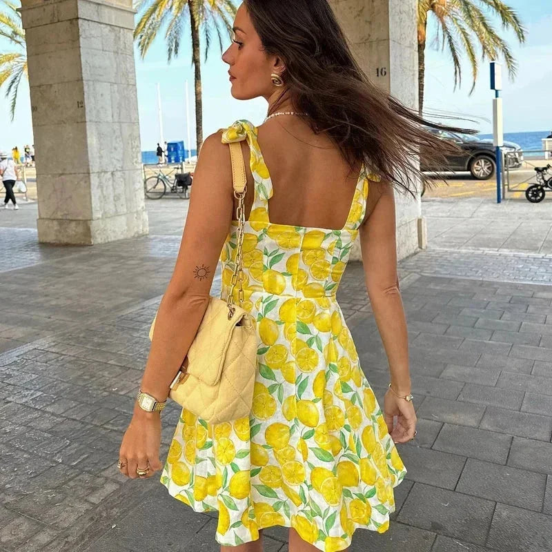 Yellow - Woman wearing a white mini dress with an all-over yellow lemon and green leaf print, featuring tie shoulder straps and a square neckline.