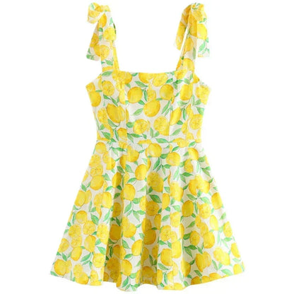 Yellow - Woman wearing a white mini dress with an all-over yellow lemon and green leaf print, featuring tie shoulder straps and a square neckline.