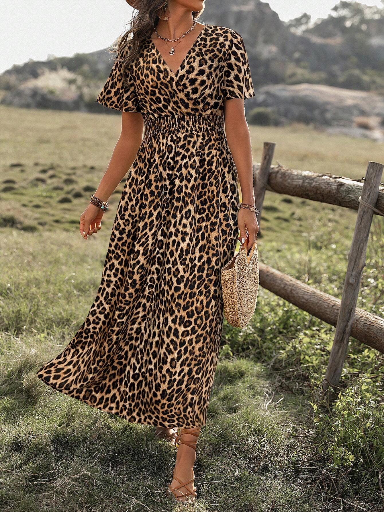 A woman wearing a leopard print maxi dress with a smocked waist, flutter sleeves, and a side slit, holding a woven straw bag in an outdoor setting.