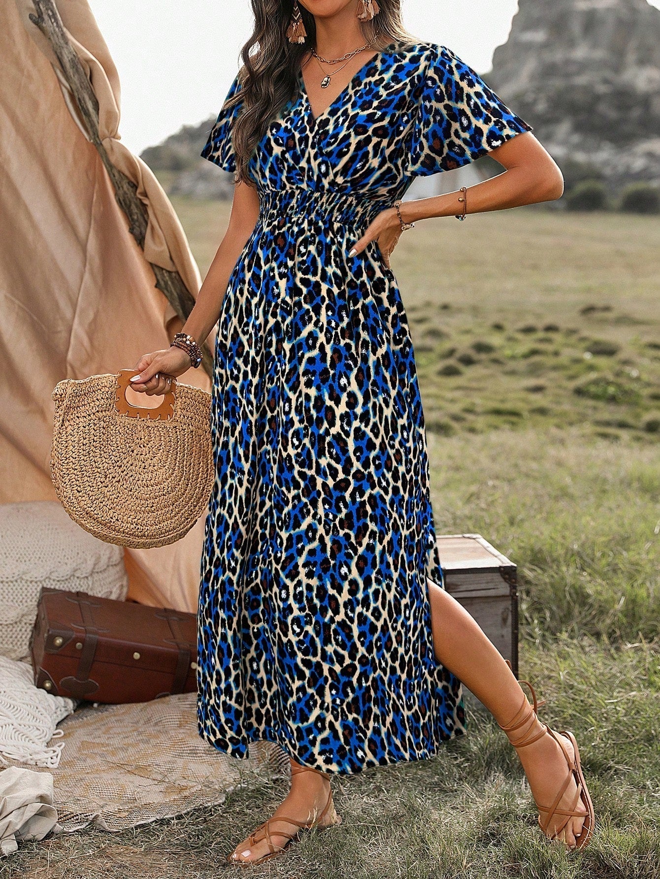 Royal Blue - A woman wearing a leopard print maxi dress with a smocked waist, flutter sleeves, and a side slit, holding a woven straw bag in an outdoor setting.