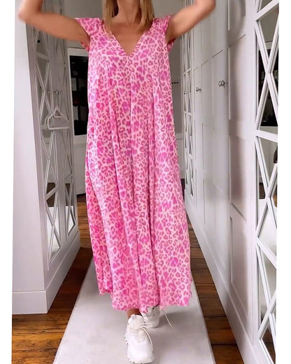 Red - A woman walks down a hallway wearing a pink leopard print maxi dress with a V-neckline and cap sleeves, carrying a woven straw tote bag.