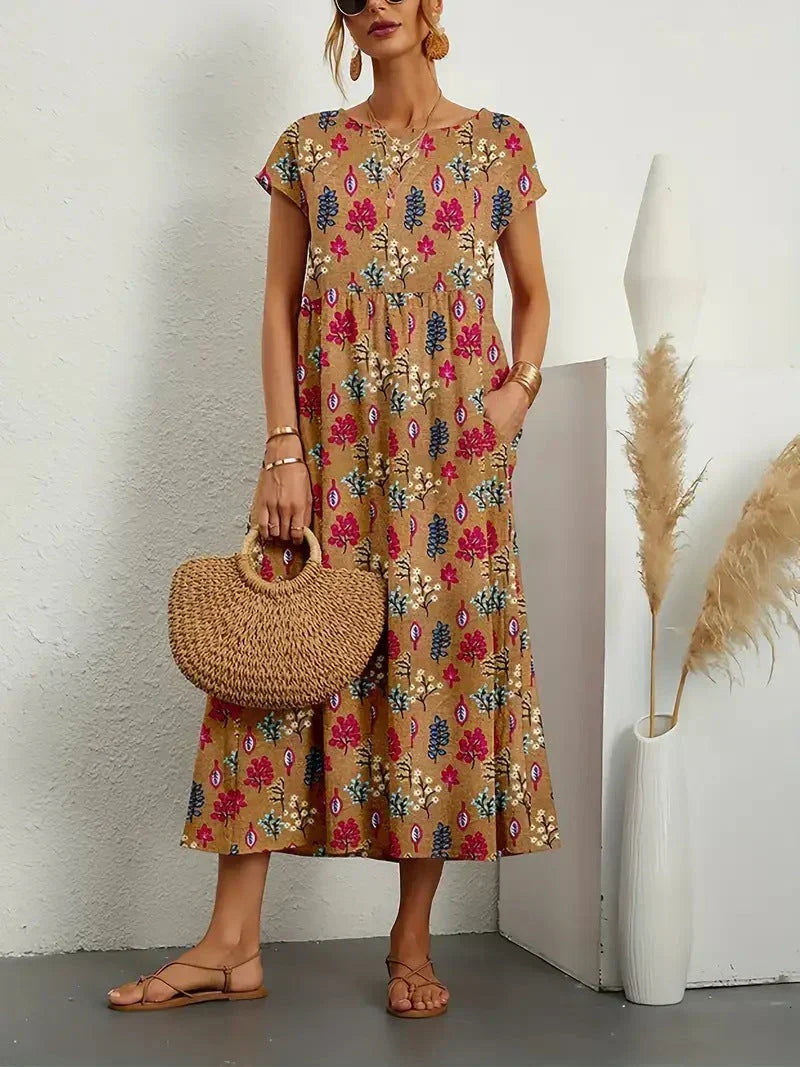Camel Garden Floral - A woman wearing a red and green kaleidoscope floral print linen-blend midi dress with cap sleeves and side pockets, holding a woven straw handbag.