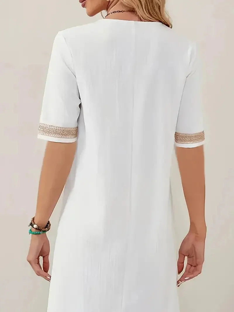 White - A woman wearing a white, knee-length shift dress made of textured linen-like fabric, featuring intricate beige crochet lace trim along the deep V-neckline and elbow-length sleeve cuffs.