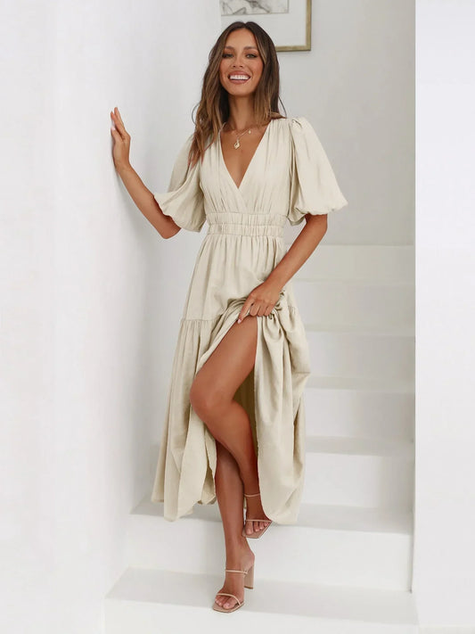 Woman wearing a beige linen maxi dress with puff sleeves, a deep V-neckline, smocked waist, and a tiered skirt with a front slit.