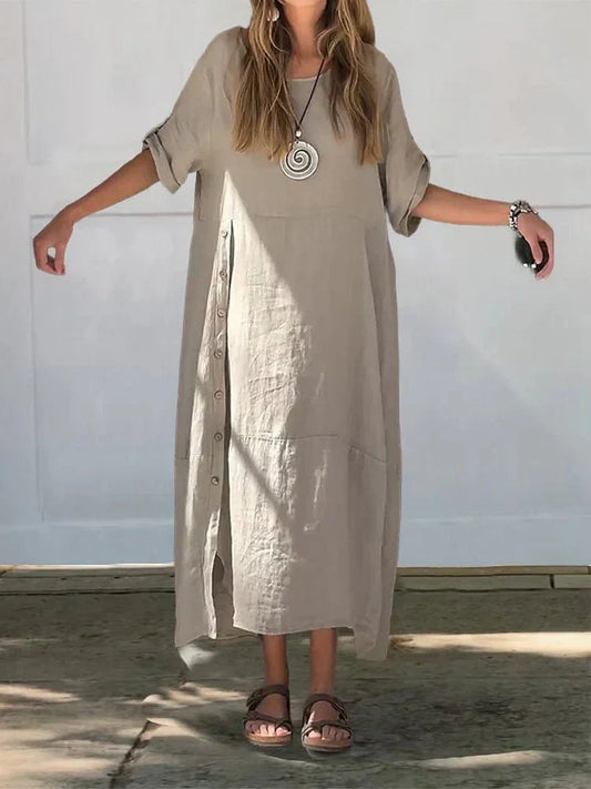 Sand - A woman wearing a loose-fitting, taupe-colored textured linen maxi dress with short cuffed sleeves, a round neck, and a decorative side button placket down the front-right side.