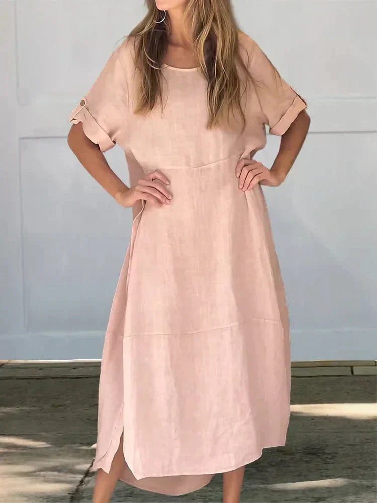 Pink - A woman wearing a loose-fitting, taupe-colored textured linen maxi dress with short cuffed sleeves, a round neck, and a decorative side button placket down the front-right side.