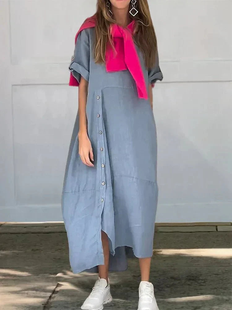 Blue - A woman wearing a loose-fitting, taupe-colored textured linen maxi dress with short cuffed sleeves, a round neck, and a decorative side button placket down the front-right side.