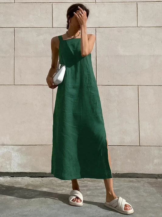 Green - Woman wearing a forest green linen-textured square neck midi dress with a side slit, styled with white sandals and a white handbag against a light stone wall.