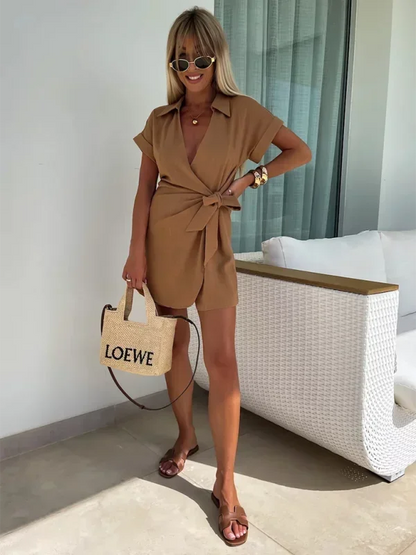 Brown - Woman wearing a rust-colored linen-blend mini dress with a collar, deep V-neck, and tie waist, styled with sandals and a straw bag.