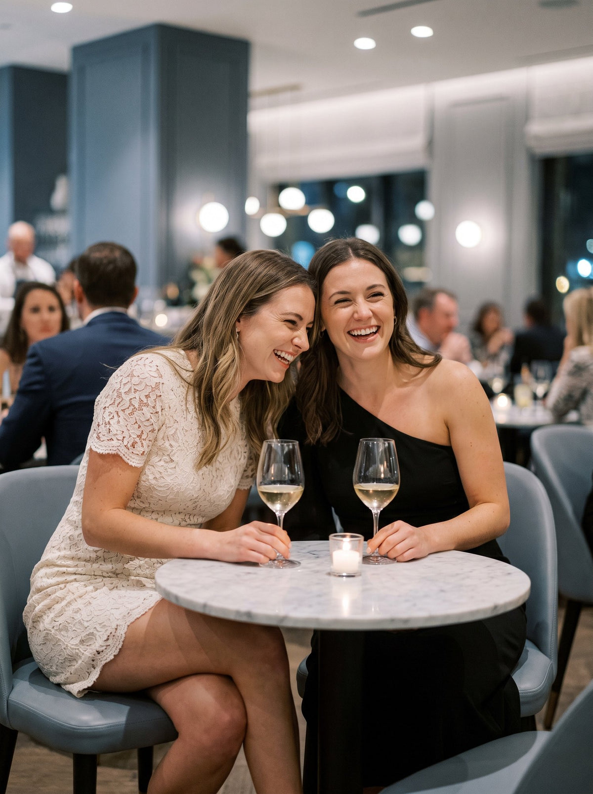 Two friends laughing in an ivory lace dress and a black one shoulder dress