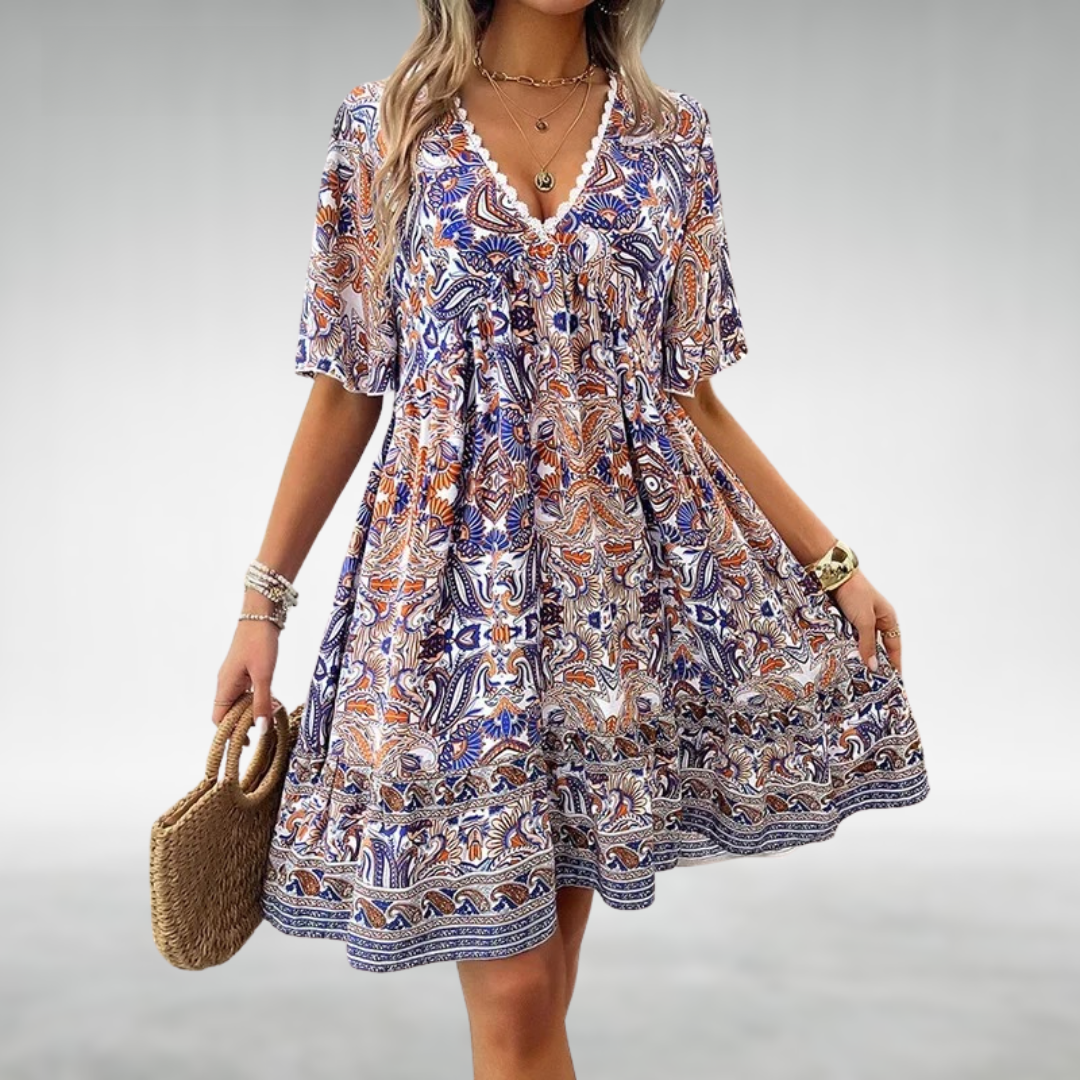 Blue - A woman wearing a bohemian-style mini dress featuring a warm-toned paisley print, a deep V-neckline with blue crochet trim, short flutter sleeves, and a tiered hem.