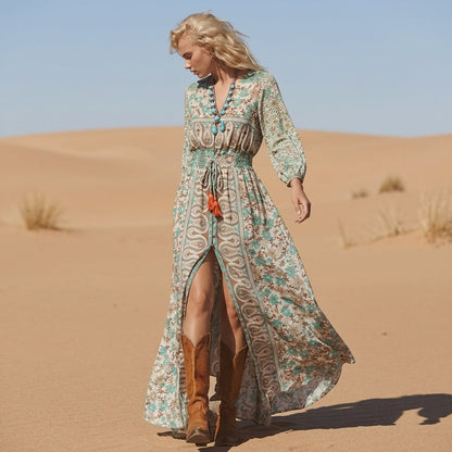 A woman stands in a desert wearing a long-sleeve, V-neck maxi dress with a green and cream paisley floral print, featuring a smocked waist, front slit, and brown leather cowboy boots.