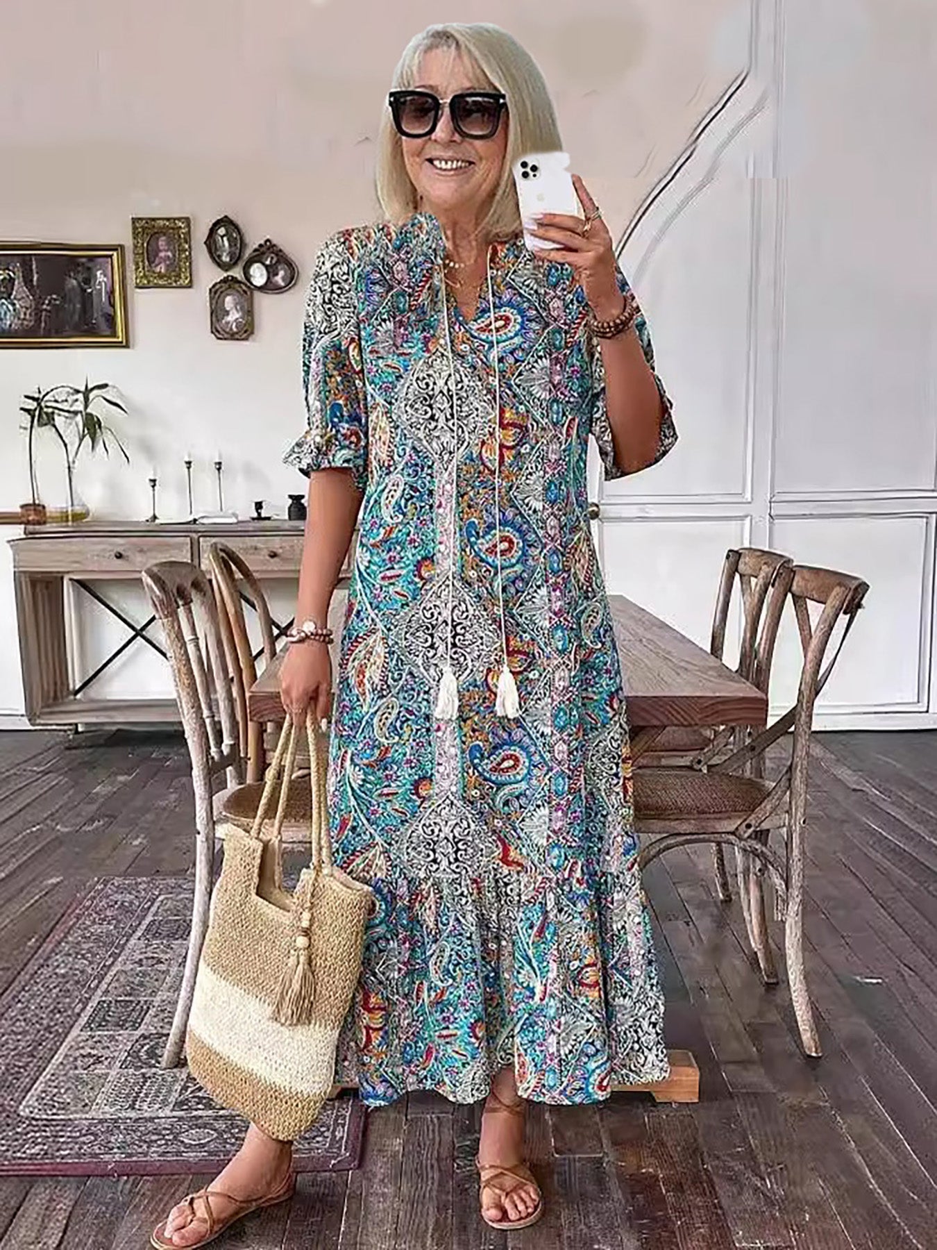 Blue - A woman wearing a colorful paisley print maxi dress with a V-neckline, tassel ties, and short sleeves, holding a woven straw tote bag.
