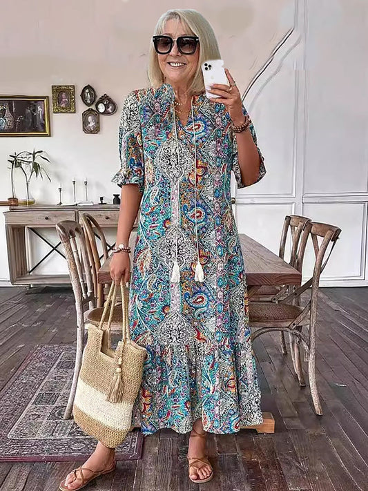 Blue - A woman wearing a colorful paisley print maxi dress with a V-neckline, tassel ties, and short sleeves, holding a woven straw tote bag.