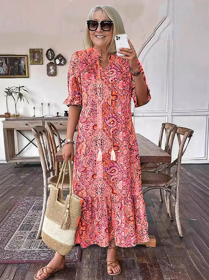 Red - A woman wearing a colorful paisley print maxi dress with a V-neckline, tassel ties, and short sleeves, holding a woven straw tote bag.