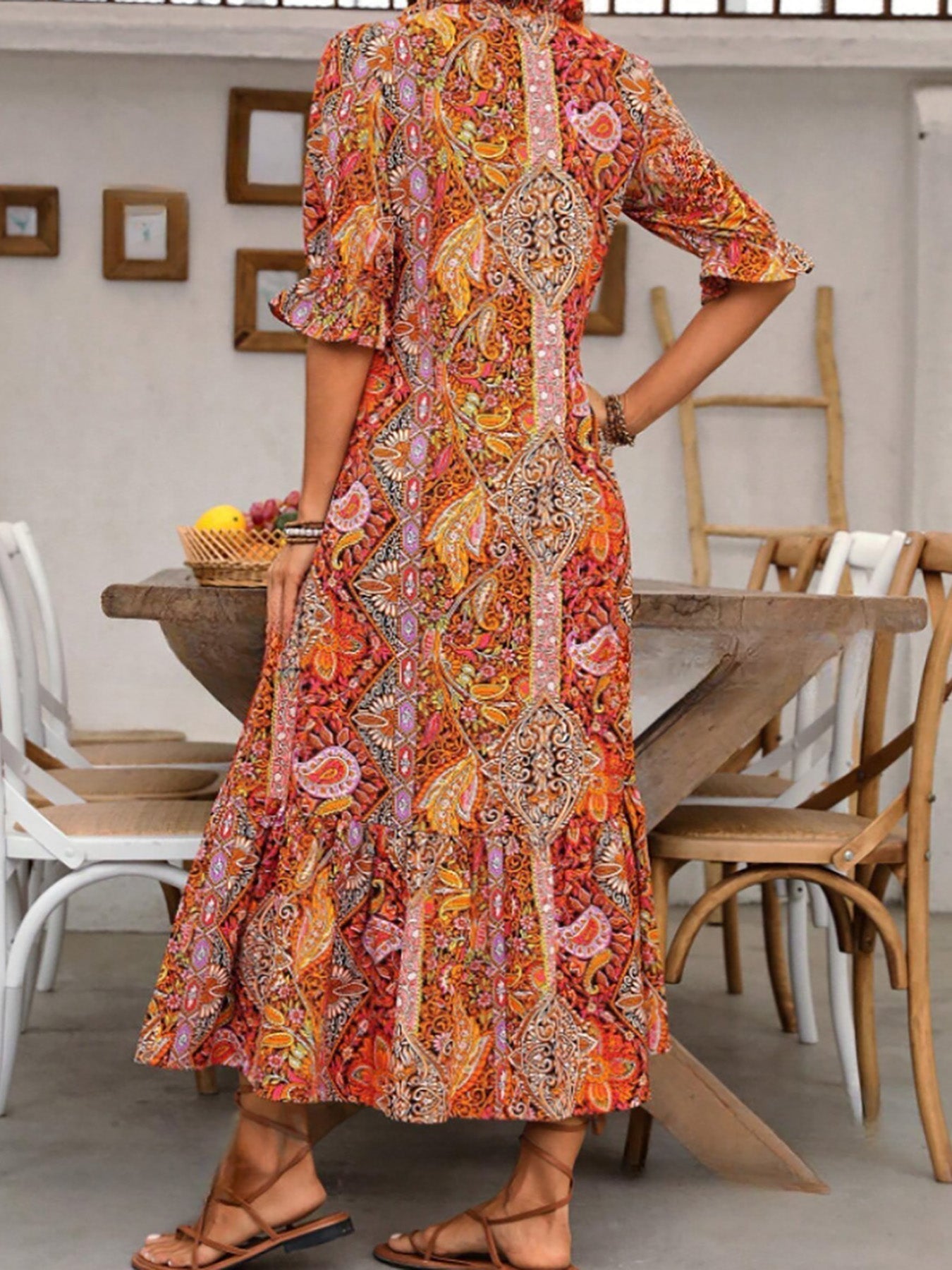 Red - A woman wearing a colorful paisley print maxi dress with a V-neckline, tassel ties, and short sleeves, holding a woven straw tote bag.