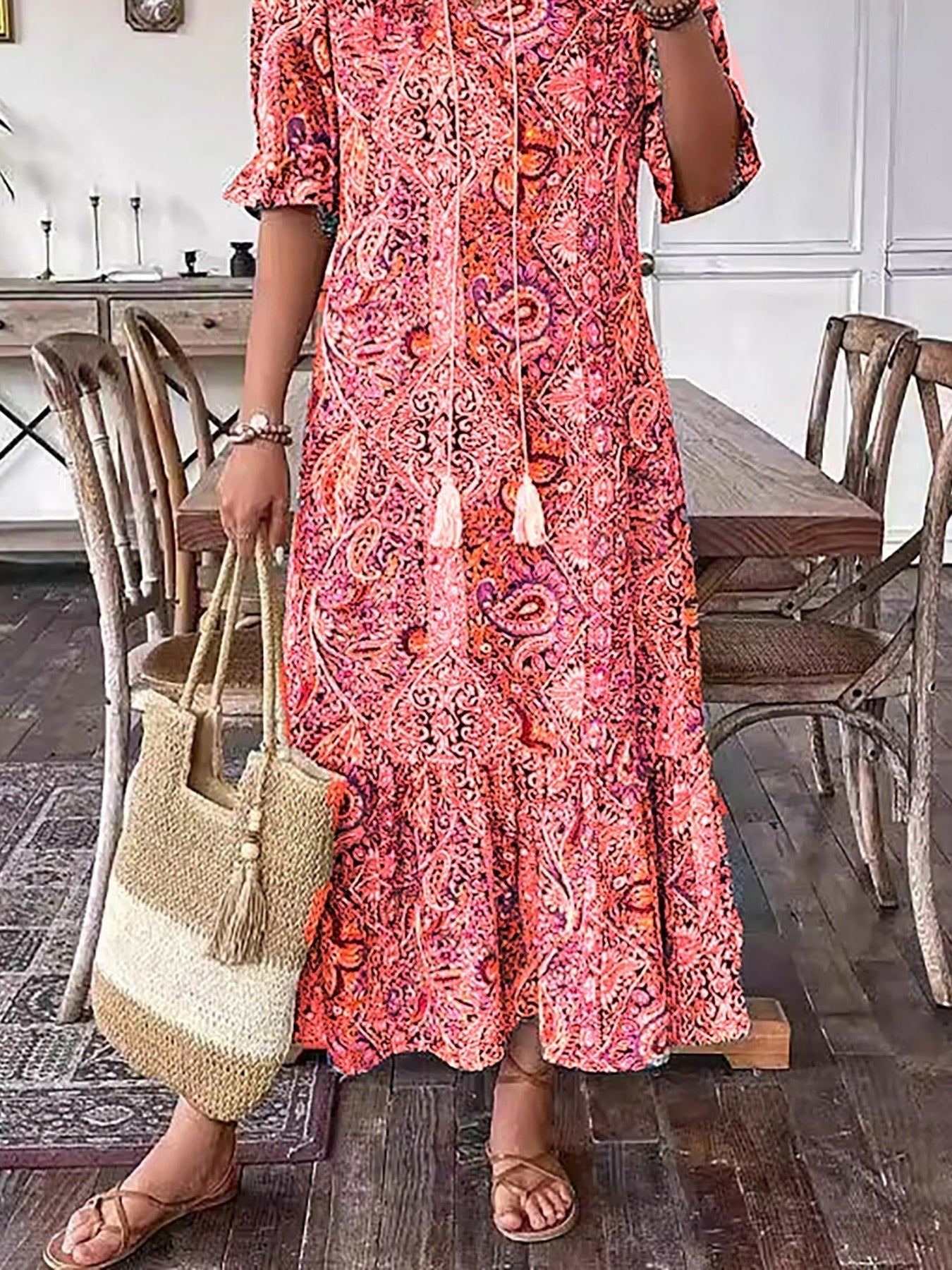 Red - A woman wearing a colorful paisley print maxi dress with a V-neckline, tassel ties, and short sleeves, holding a woven straw tote bag.