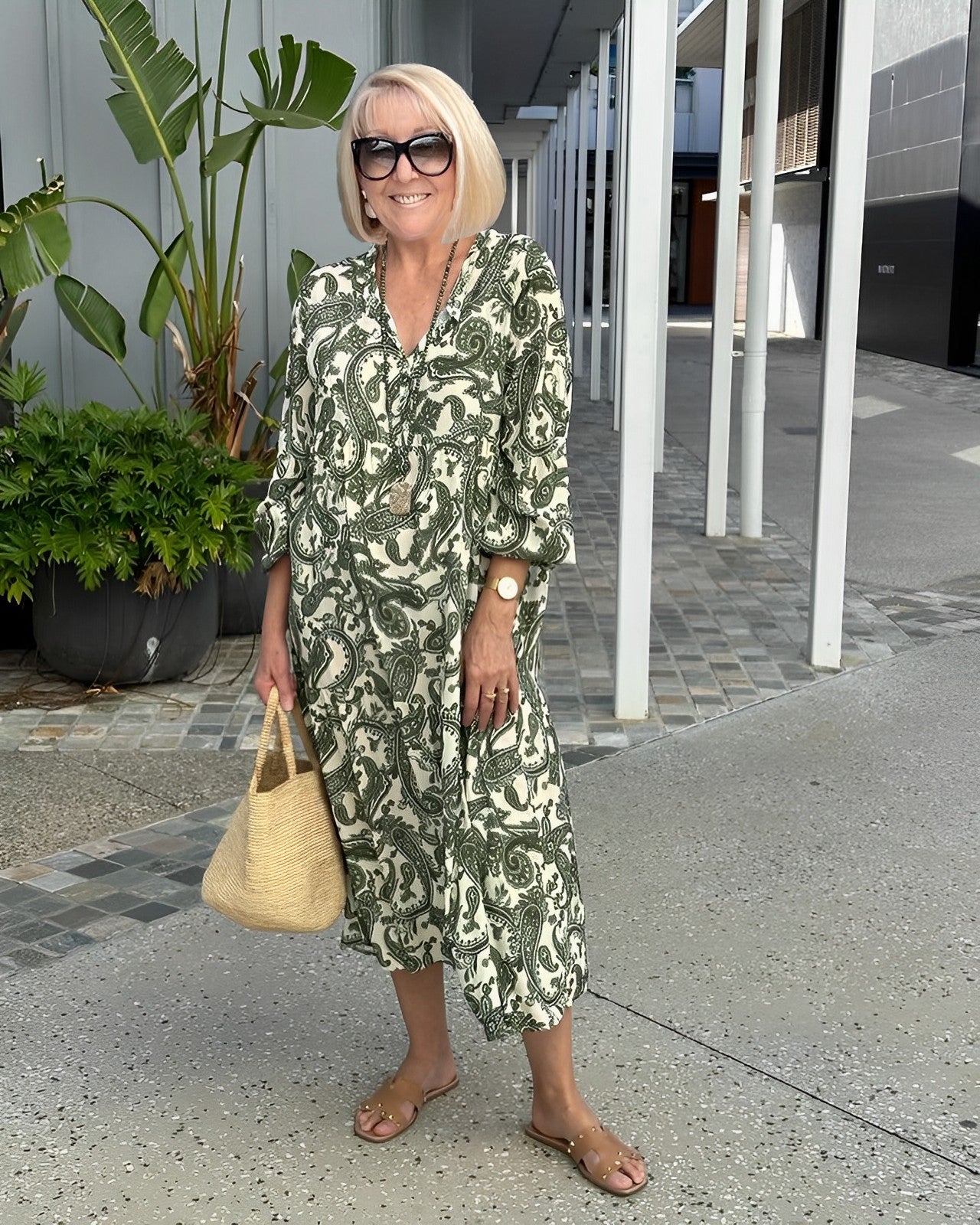 A woman wearing a green and cream paisley print midi dress with balloon sleeves, a v-neckline, paired with brown slide sandals and a woven straw bag.