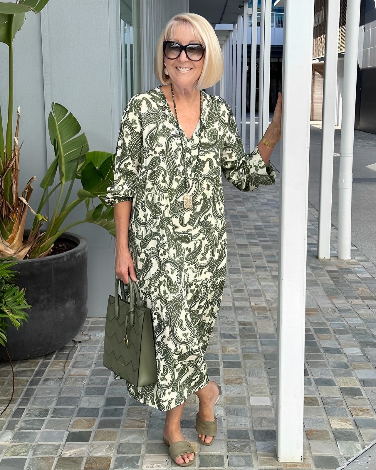 A woman wearing a green and cream paisley print midi dress with balloon sleeves, a v-neckline, paired with brown slide sandals and a woven straw bag.