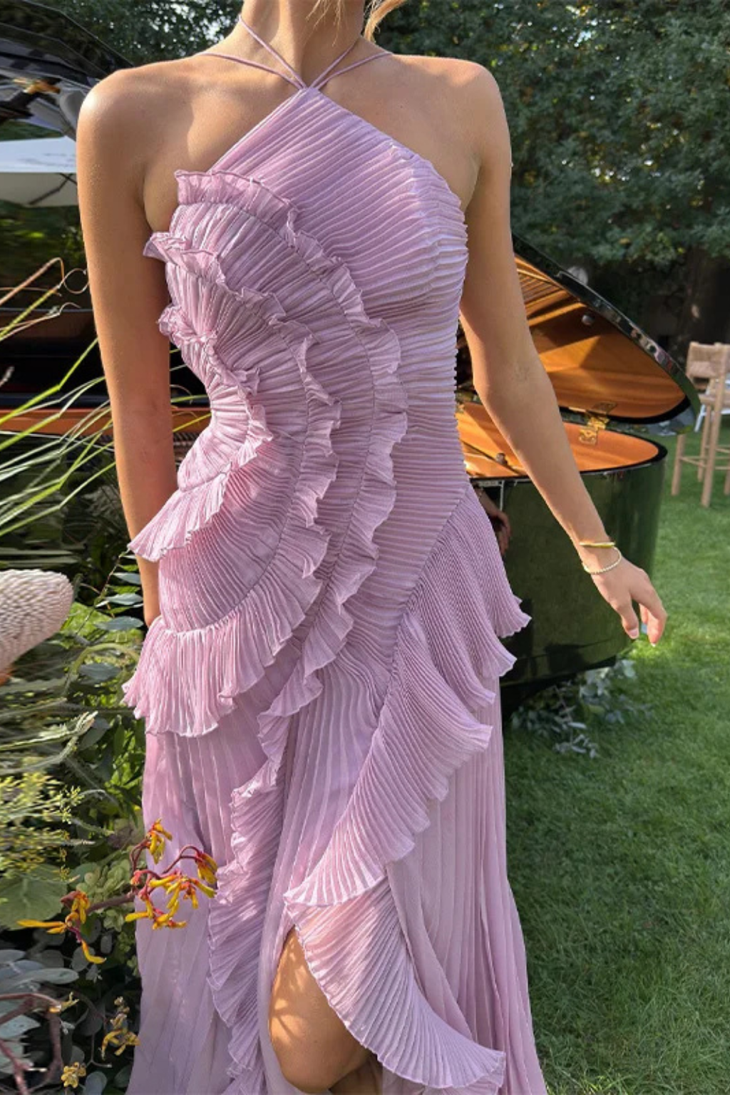 Purple - A woman wearing a sleeveless, light pink halter maxi dress made of pleated chiffon, featuring a cascading ruffle down the front and an asymmetrical high-low hemline.