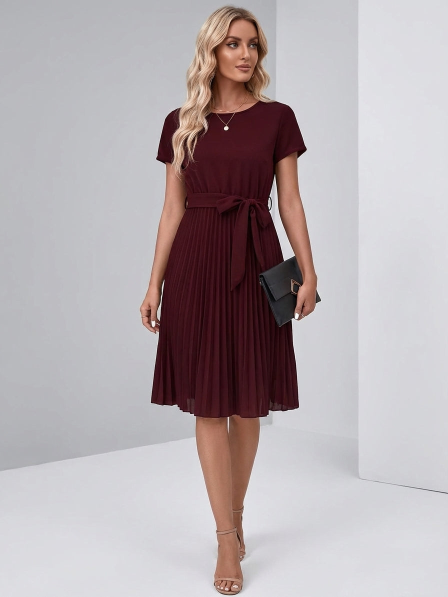 Purple - A woman wearing a short-sleeve, crew-neck burgundy midi dress with a belted waist and a pleated accordion skirt, holding a black clutch.
