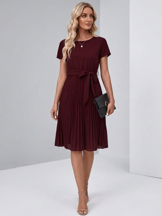 Purple - A woman wearing a short-sleeve, crew-neck burgundy midi dress with a belted waist and a pleated accordion skirt, holding a black clutch.