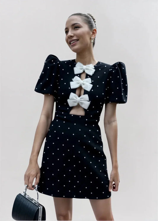 Black mini dress with white polka dots, featuring short puff sleeves, a center front cutout, and three contrasting white 3D bows down the bodice.