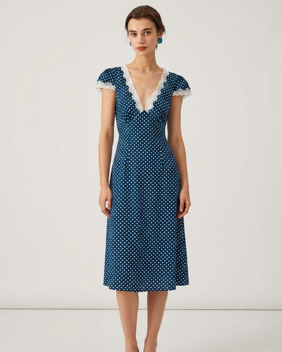 Navy - A woman wearing a navy blue and white polka dot midi dress with white lace trim along the V-neckline and cap sleeves, paired with white low-heeled slide sandals.