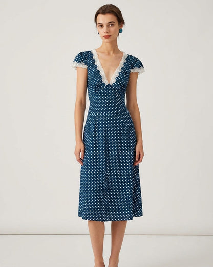 Navy - A woman wearing a navy blue and white polka dot midi dress with white lace trim along the V-neckline and cap sleeves, paired with white low-heeled slide sandals.