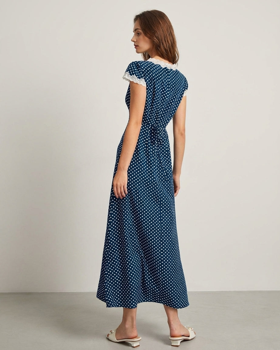 Navy - A woman wearing a navy blue and white polka dot midi dress with white lace trim along the V-neckline and cap sleeves, paired with white low-heeled slide sandals.