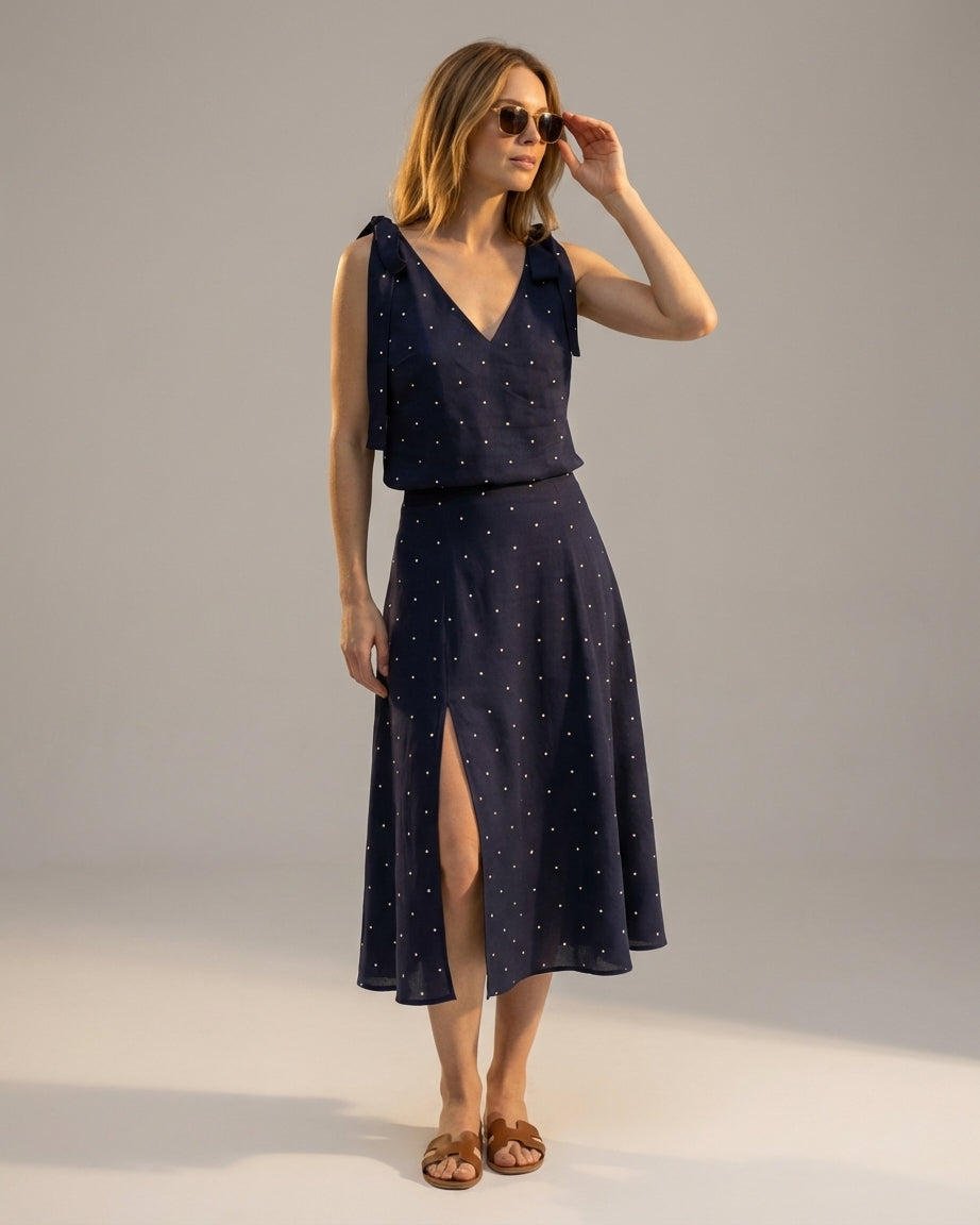 Navy - A woman wearing a navy blue and white polka dot two-piece midi set, featuring a cropped tie-shoulder V-neck top and a high-waisted midi skirt with a side slit.
