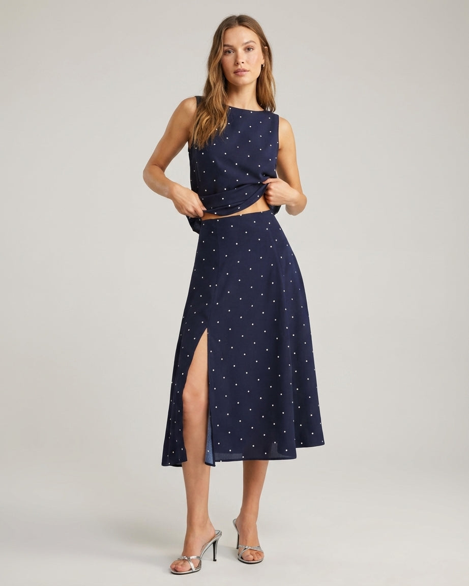 Navy - A woman wearing a navy blue and white polka dot two-piece midi set, featuring a cropped tie-shoulder V-neck top and a high-waisted midi skirt with a side slit.