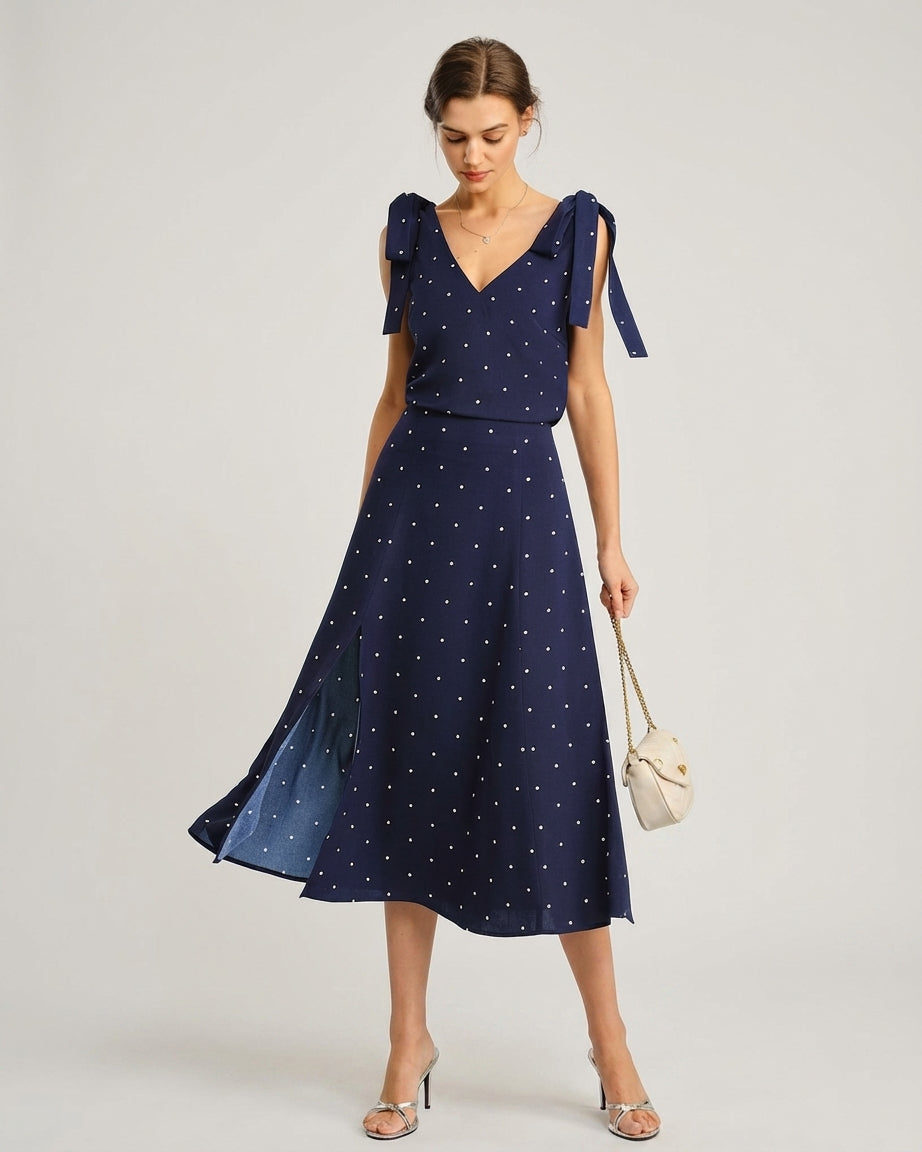 Navy - A woman wearing a navy blue and white polka dot two-piece midi set, featuring a cropped tie-shoulder V-neck top and a high-waisted midi skirt with a side slit.
