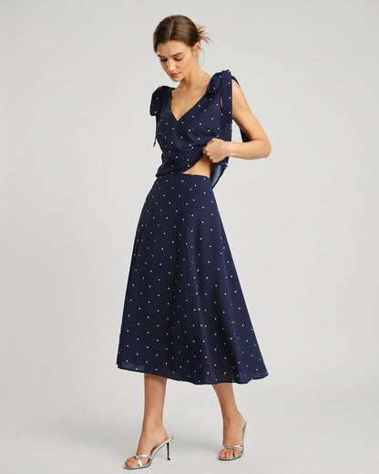 Navy - A woman wearing a navy blue and white polka dot two-piece midi set, featuring a cropped tie-shoulder V-neck top and a high-waisted midi skirt with a side slit.