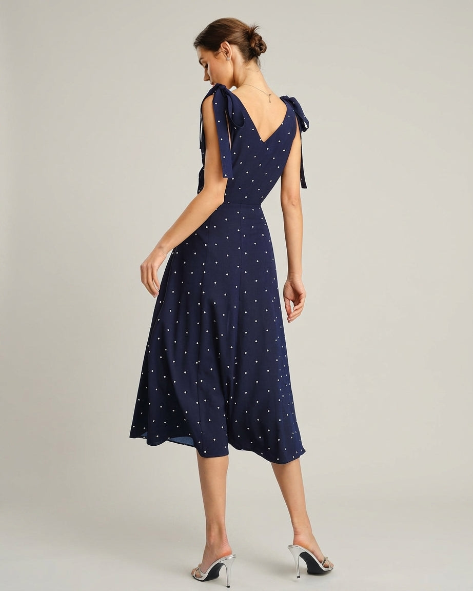 Navy - A woman wearing a navy blue and white polka dot two-piece midi set, featuring a cropped tie-shoulder V-neck top and a high-waisted midi skirt with a side slit.