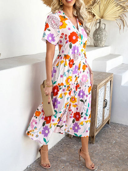 Blush Pink - A woman wearing a short-sleeve, collared maxi dress with a colorful retro floral print, featuring a button-down front and a tiered skirt.
