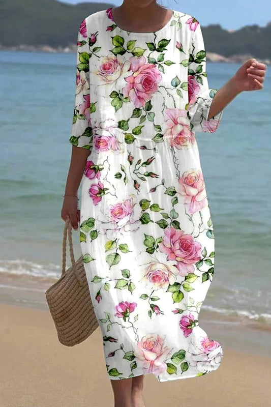 Floral - A woman on a beach wearing a white midi dress featuring a pink rose and green leaf print, three-quarter sleeves, and a relaxed linen-texture drape.