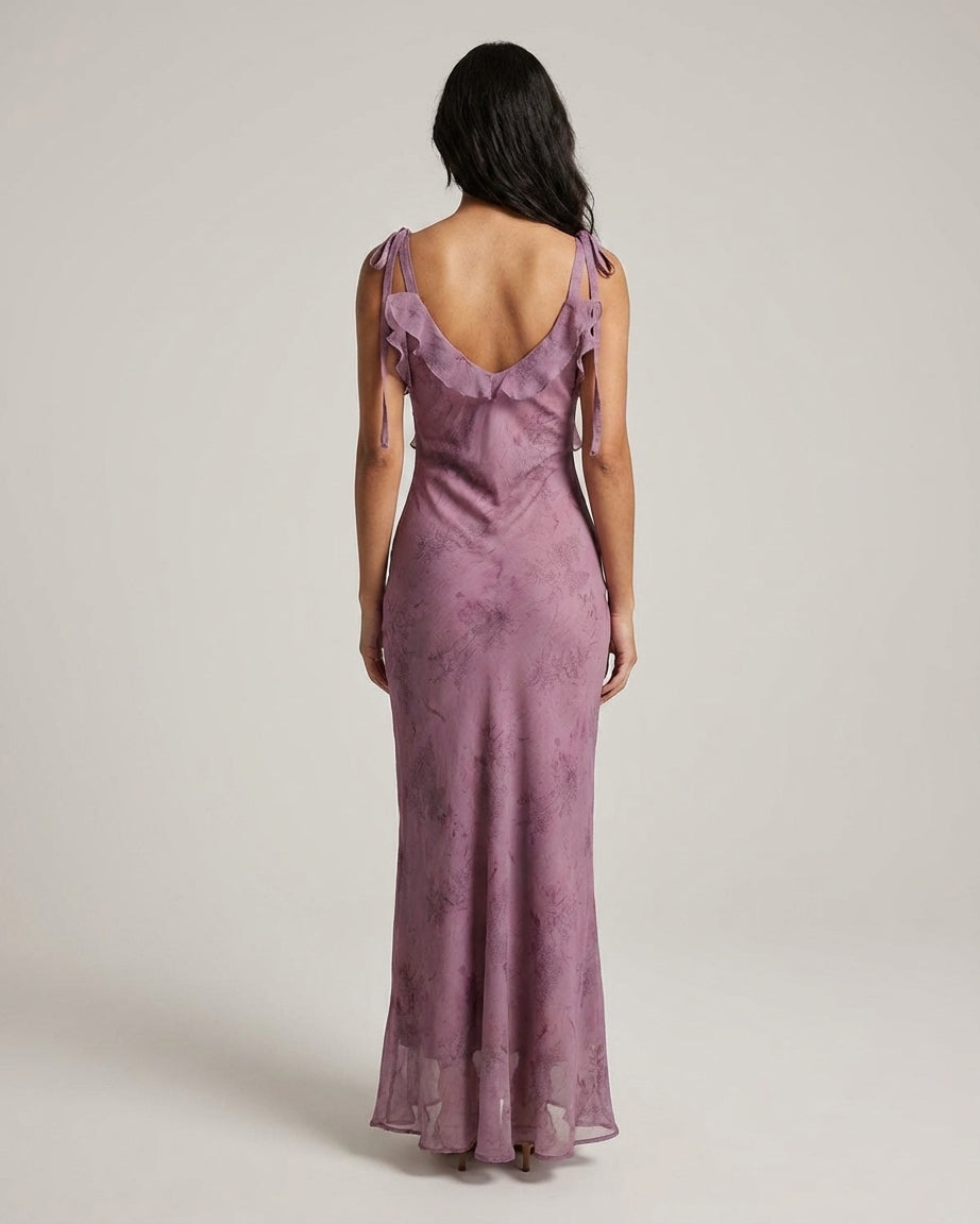 Purple - A woman wearing a dusty purple midi dress with a dark abstract floral print, featuring adjustable tie straps, a ruffled V-neckline, and a sheer chiffon hem.