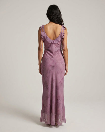 Purple - A woman wearing a dusty purple midi dress with a dark abstract floral print, featuring adjustable tie straps, a ruffled V-neckline, and a sheer chiffon hem.