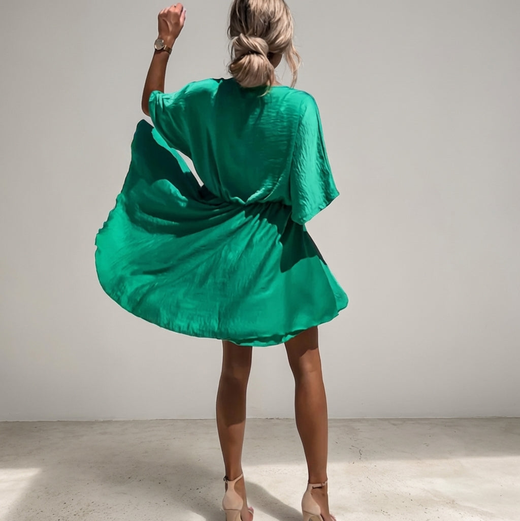 Dark Green - A woman wearing a light olive green satin mini dress featuring a deep V-neck, twist-front waist detail, short flutter sleeves, and a tiered asymmetric skirt, paired with nude heeled sandals.