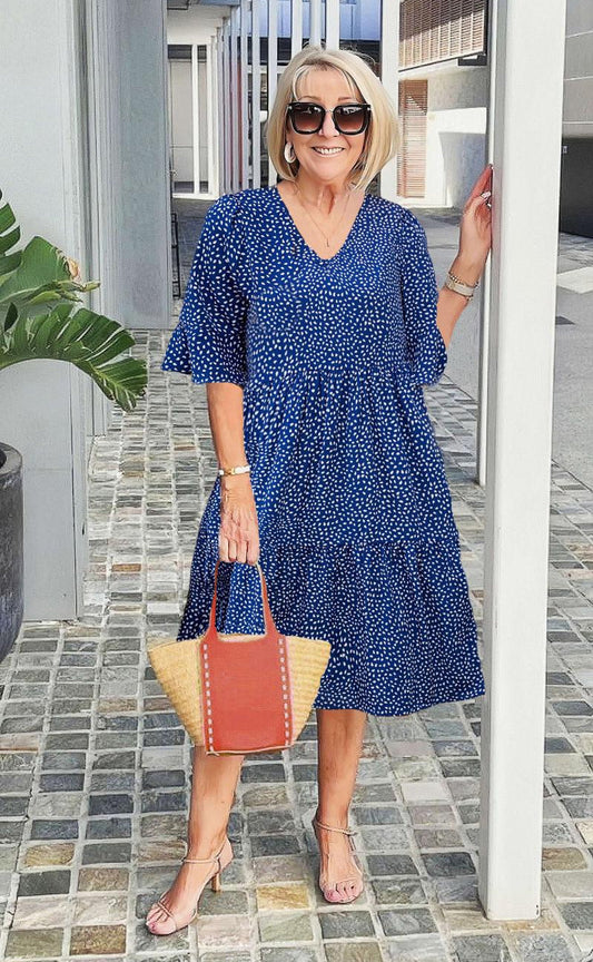 A woman wearing a blue and white speckled print midi dress with a V-neckline, elbow-length flutter sleeves, and a tiered skirt, holding a woven straw tote bag.