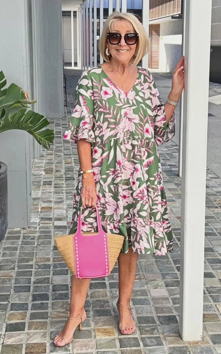 A woman wearing a blue and white speckled print midi dress with a V-neckline, elbow-length flutter sleeves, and a tiered skirt, holding a woven straw tote bag.